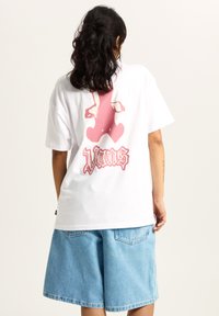 Hvid bomulds t-shirt med et pink grafisk bjørnedesign på ryggen og "Vans" tekst nedenunder. Sammen med lyseblå denim shorts.