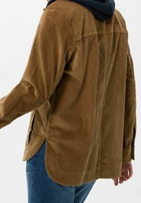 Camicia in corduroy marrone con vestibilità rilassata, caratterizzata da una texture uniforme, maniche lunghe, aperture laterali e orlo curvo. Vista posteriore mostrata.