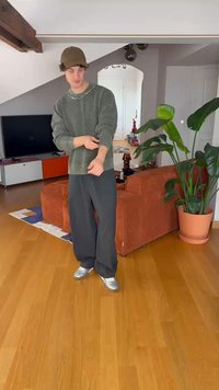 Přerostlý zelený pletený svetr, šedé kalhoty s širokými nohavicemi a stříbrné tenisky. Neformální vnitřní prostředí s dřevěnou podlahou a květináčem.