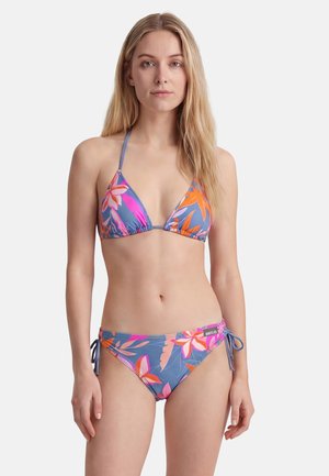Junge Frau trägt einen blauen Bikini mit pinken und orangefarbenen Blumenmustern und steht vor einem schlichten weißen Hintergrund.