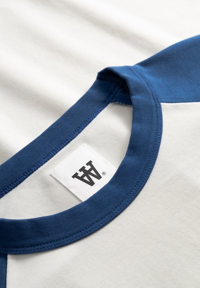 Λευκό βαμβακερό t-shirt με σκούρο μπλε ριμπ λαιμόκοψη και λεπτομέρειες στα μανίκια. Διαθέτει τυπωμένη ετικέτα στο εσωτερικό του γιακά.