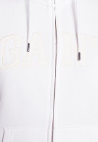 White zip-up hoodie made of soft fabric, featuring embossed "GANT" logo in cream, silver zipper hardware, and drawstring at the hood.