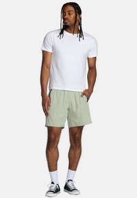 Shorts de bain vert clair avec une taille élastique, associés à un t-shirt blanc ajusté et des baskets noires avec des accents blancs.