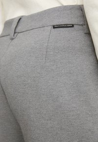 Gros plan sur un pantalon en tissu gris avec une poche latérale et une étiquette noire "Marc O'Polo Denim" près de la taille, associé à un haut blanc cassé.