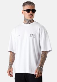 Mann mit Nacken- und Armtattoos, der schwarze Sonnenbrillen trägt, ein übergroßes weißes T-Shirt mit kleinen schwarzen Schriftzügen und schwarze Hosen.