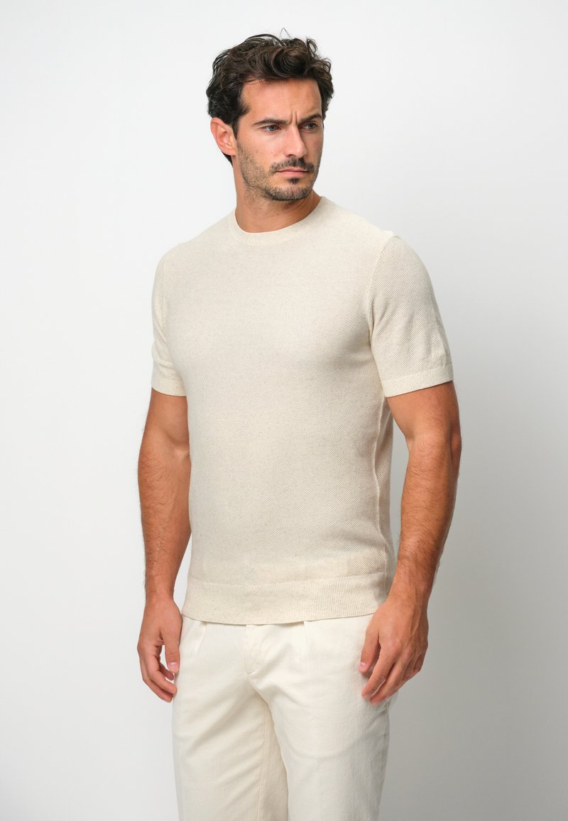 Beige Kurzarm-Strickpullover mit rundem Ausschnitt, der ein dezentes Strukturmuster und eine figurbetonte Form aufweist. Kombination mit hellen Hosen.