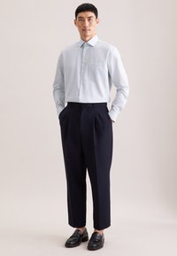 Hellblaues Hemd mit Knöpfen und kleinem Muster, kombiniert mit marineblauen, plissierten Hosen und schwarzen Loafers, stehend vor neutralem Hintergrund.