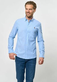 Világoskék, gombos ing, amely pamutból készült, klasszikus gallérral és hosszú ujjaival, gombos mandzsettával. A mellkason egy kis logó díszíti.