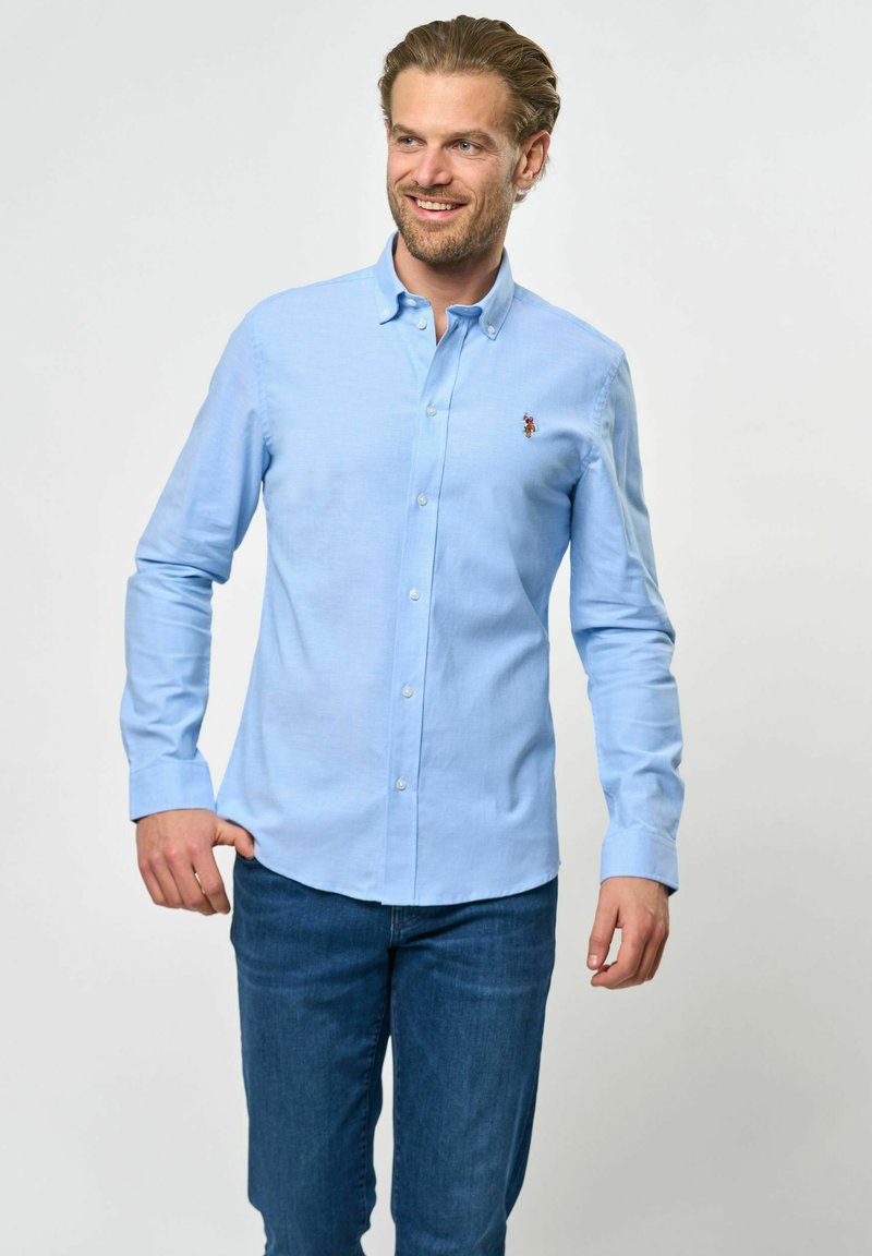 Világoskék, gombos ing, amely pamutból készült, klasszikus gallérral és hosszú ujjaival, gombos mandzsettával. A mellkason egy kis logó díszíti.