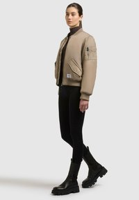 Beige Bomberjacke mit gerippten Bündchen und Saum, mit Reißverschlusstasche am Ärmel, kombiniert mit schwarzen Leggings und klobigen schwarzen Stiefeln.