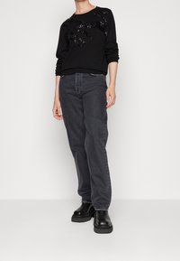 Svart sweatshirt med paljettdetalj på framsidan, kombinerad med mörkgrå jeans och svarta kängor med tjock sula. Enfärgad bakgrund.