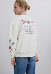 Kermanvärinen collegepaita, jossa on painettuja tekstejä ja tähtiä punaisina, sinisinä ja hopeisina takana ja hihassa. Rentoa mallia, resorireunuksilla.