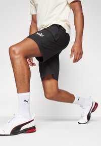 Pantalones cortos deportivos negros con bolsillos, logo de Puma en blanco, combinados con calcetines blancos con un logo en negro y zapatillas blancas con detalles rojos.