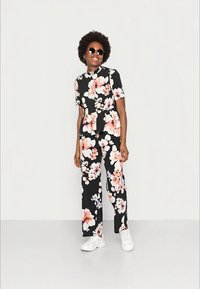 Macacão preto floral com mangas curtas e calças largas, adornado com padrões de flores rosas e brancas. Combinado com ténis brancos.