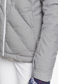 Graue, gepolsterte Jacke mit diagonaler Naht, weißem Reißverschluss und einer Seitentasche. Der Ärmel zeigt einen gemusterten Stoff darunter.