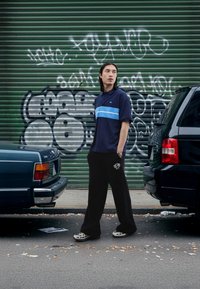 Marinblå pikétröja med blå rand, lösa svarta byxor och mönstrade skor. Urban bakgrund med graffiti och parkerade fordon.