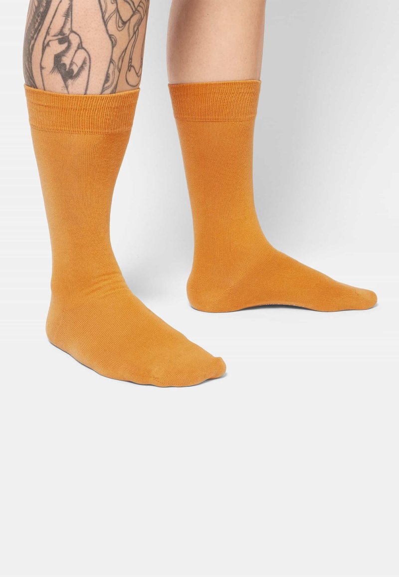 Oranje katoenen sokken met geribbelde boorden. Glad textuur en crewlengte, gedragen op voeten met een getatoeëerd been.