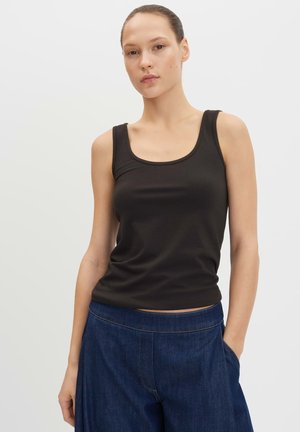 Donna che indossa un top nero aderente senza maniche e pantaloni di denim blu scuro larghi con una mano in tasca su sfondo semplice.