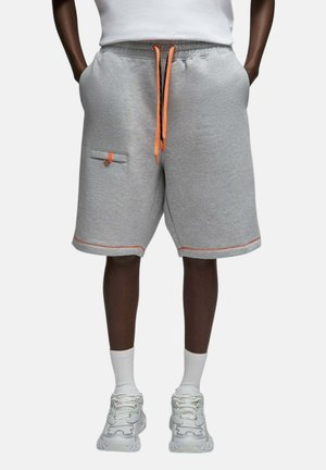 Personne portant un short gris à cordon avec des accents orange, des chaussettes blanches et des baskets blanches, les mains dans les poches, debout devant un fond uni.