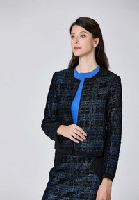 Veste texturée noire et bleue au design court, avec un col rond et des accents noirs, associée à un haut bleu. Tissu lisse avec une surface à motifs.
