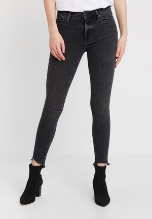 Frau trägt hoch taillierte schwarze Skinny-Jeans mit offenem Saum und schwarze Stiefeletten mit Absatz vor schlichtem Hintergrund.