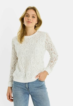 Mujer con cabello rubio largo, que lleva pendientes de aro dorados, una blusa blanca de encaje de manga larga y pantalones vaqueros azul claro, de pie con una mano en el bolsillo.