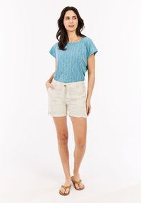 Lichtblauw patroon top met korte mouwen, gecombineerd met beige shorts met zijzakken en een verstelbare taille. Bruine flip-flops.