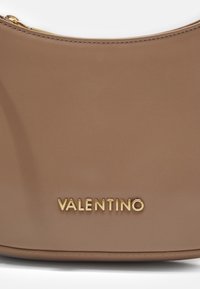 Tuvumā redzams beidzējo Valentino rokassomu, kas izgatavota no bežīgas ādas ar zelta rāvējslēdzēju un zelta Valentino logo uz gludas ādas virsmas.