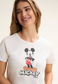 Camiseta de algodón blanca con un gráfico de Mickey Mouse en estilo de dibujo animado en negro, rojo y naranja. Texto "MICKEY" en letras negras en negrita debajo del gráfico.