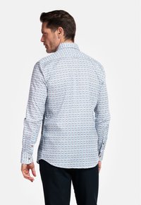 Chemise blanche à manches longues avec un motif géométrique bleu et orange, boutons de poignet, et col classique, portée avec un pantalon foncé.