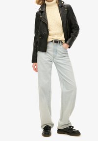 Giacca in pelle nera da motociclista, maglione beige a treccia, jeans a gamba larga blu chiaro e scarpe nere con dettagli a frange. Look semplice e informale.