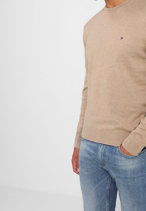 Homme portant un pull beige à col rond avec un petit logo sur la poitrine et un jean bleu clair, main dans la poche devant un fond uni.
