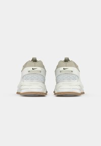 Par de sapatilhas Nike Zoom em bege e branco vistas de trás, com design geométrico no calcanhar e solas de borracha castanha, sobre fundo branco.