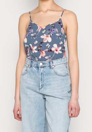 Kvinde iført blå blomstrede spaghetti-stropbluse stoppet ned i højtaljede jeans i lys vask mod en ensartet baggrund.