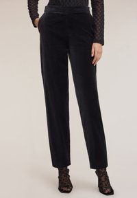 Kobieta w czarnych, welurowych spodniach z wysokim stanem i bocznymi kieszeniami, w połączeniu z przezroczystymi, czarnymi skarpetkami w panterkę oraz czarnymi sandałkami na obcasie.