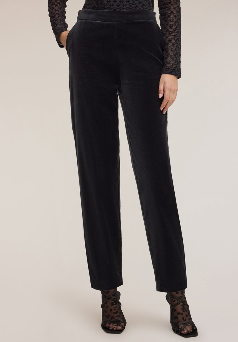 Kobieta w czarnych, welurowych spodniach z wysokim stanem i bocznymi kieszeniami, w połączeniu z przezroczystymi, czarnymi skarpetkami w panterkę oraz czarnymi sandałkami na obcasie.