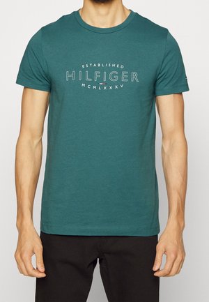 Mann trägt ein türkisfarbenes Kurzarm-T-Shirt mit dem Text "Established Hilfiger MCMLXXXV" und kleinem Logo auf dem linken Ärmel, kombiniert mit schwarzen Hosen.