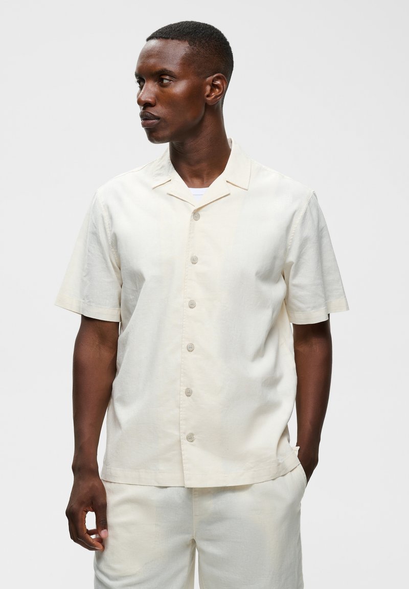 Homme aux cheveux courts portant une chemise crème à manches courtes boutonnée et un pantalon assorti, regardant vers la gauche avec une main dans la poche.