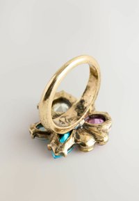 Auksinės spalvos žiedas su lygiu, plonu žiedu, įrėmintu gėliniu pagrindu, papuoštu mėlynais, rožiniais ir skaidriais kristalų akcentais įvairiomis formomis.