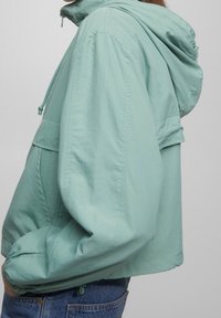 Ljus teal, kort jacka med huva, som har elastiska ärmslut och en framficka. Tillverkad av ett mjukt, lätt material.