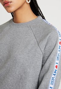 Felpa grigia con collo a girocollo a costine, caratterizzata da testo blu e rosso "I ♥ MY VANS" lungo la manica. Il tessuto appare morbido e strutturato.
