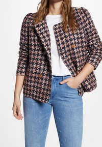 Blazer de cuadro de perro de doble botonadura en tonos azul marino, rosa y crema; tejido texturizado con botones marrones; llevado sobre una blusa blanca y jeans de color azul claro.