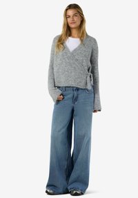 Ung kvinna klädd i en grå omslagströja, vit skjorta, vida blå jeans och silverfärgade sneakers, stående mot en vit bakgrund.