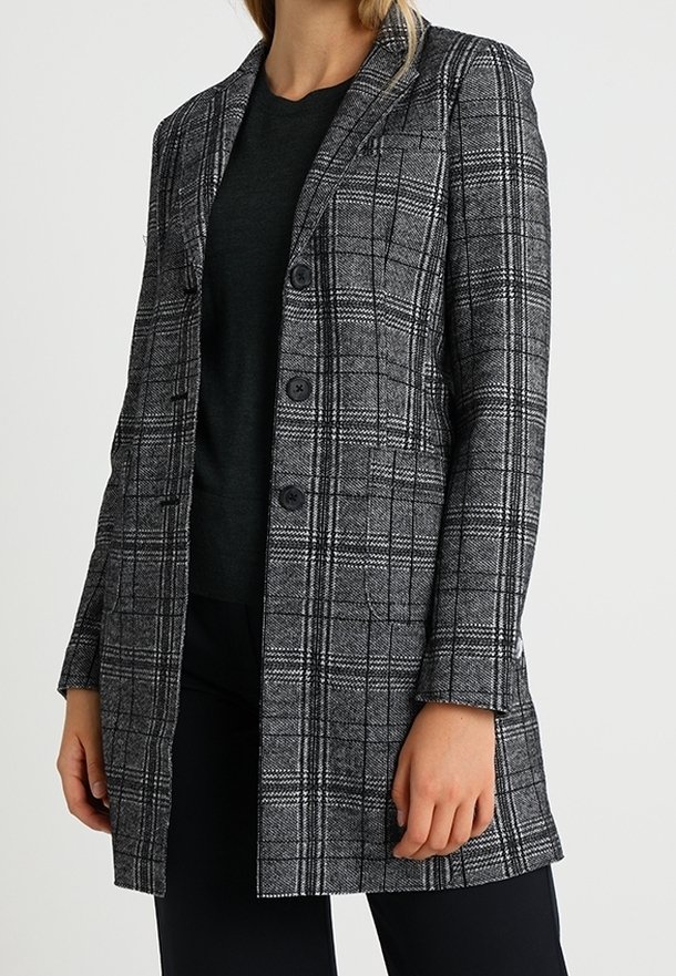 Femme portant un long blazer à carreaux gris et noir par-dessus un haut noir et un pantalon noir, debout avec sa main gauche détendue le long du corps.