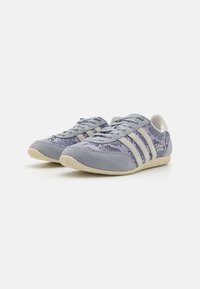 Scarpe sportive grigio chiaro con una tomaia testurizzata, caratterizzate da accenti viola e tre strisce bianche. Suola in gomma con un design a profilo basso.