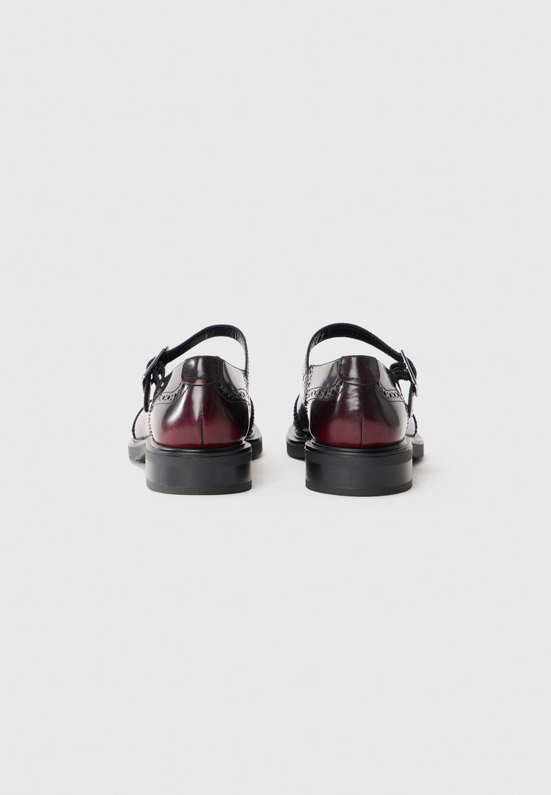 Chaussures en cuir noir avec une finition brillante, dotées d'un talon surélevé accentué de rouge, d'une sangle réglable à la cheville et de détails décoratifs perforés.