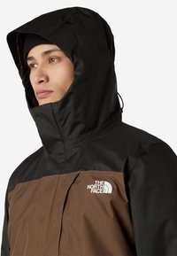 Kurtka z kapturem z czarną górą i brązowym dołem, wykonana z teksturowego materiału. Logo na klatce piersiowej, elegancki fason z zapięciem na zamek.