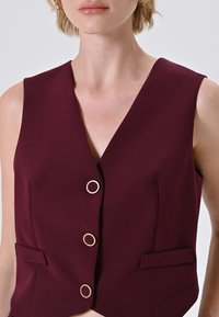 Gilet sans maniche bordeaux realizzato in tessuto liscio, con profondo scollo a V, tre bottoni dorati e due tasche frontali.