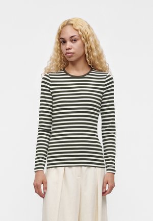 Jeune femme aux longs cheveux blonds bouclés portant un haut à manches longues rayé vert foncé et blanc et un pantalon large crème.