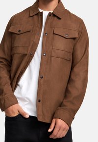 Homme portant une veste à boutons marron avec des poches à rabat sur un t-shirt blanc et un pantalon noir, la main dans la poche, contre un fond uni.